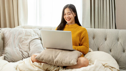An attractive Asian woman in comfy clothes is using a laptop computer on a couch at home.