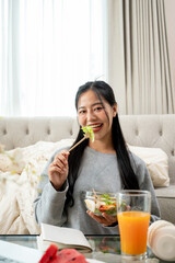 Fototapeta premium A charming Asian woman is enjoying eating healthy salad at a coffee table in the living room.