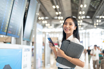 A beautiful Asian woman with her passport is standing in front of a flight information display.