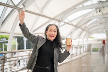 A cheerful mature Asian businesswoman is waving her hand at the camera while standing on a skywalk.