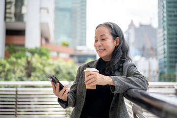 A happy mature Asian businesswoman is reading messages on her phone while standing on a skywalk.