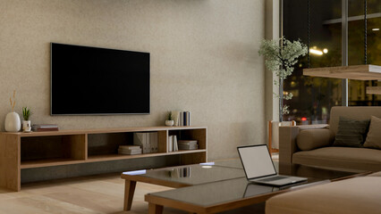 Contemporary living room at night featuring a laptop on a coffee table, a sofa, and a TV on the wall © bongkarn