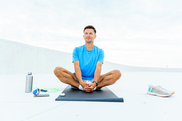 A man sits cross-legged on a mat, embracing a tranquil moment after exercise, surrounded by fitness...
