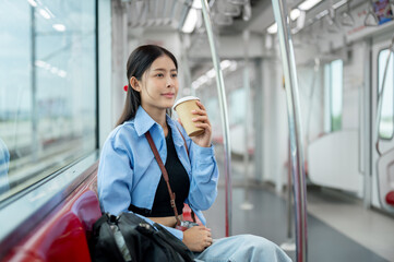 A happy Asian female passenger is sipping coffee while commuting in the city with a sky train.