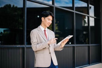 Fototapeta premium Confident Asian man with a smile standing holding notepad and tablet at out side office. big city on business center
