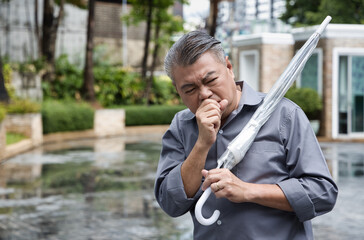 Ill Asian middle-aged man carrying umbrella after raining; sick Asian uncle coughing; sore throat;...