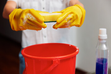 Woman in yellow rubber gloves holding a sponge over a red bucket with a bottle of cleaning solution