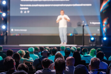 Speaker on stage delivering a presentation to a diverse audience at a professional conference. Engaging and informative public speaking event.