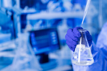 A person is holding a beaker with a liquid in it. The background has a computer monitor and a keyboard. Scene is scientific and focused