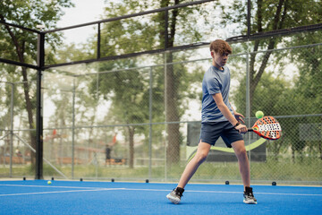 Focused young male player hitting the ball during a game of padel tennis on an outdoor blue court.