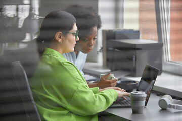 Shot through office window blinds of female African American software developer supervising...