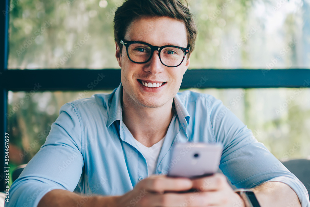 Poster portrait of cheerful male blogger smiling at camera while messaging in online chat with friend on mo