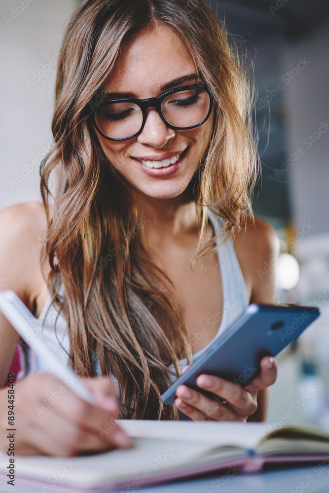Poster positive young woman in eyeglasses making notes in notepad sitting at table and using free wifi conn