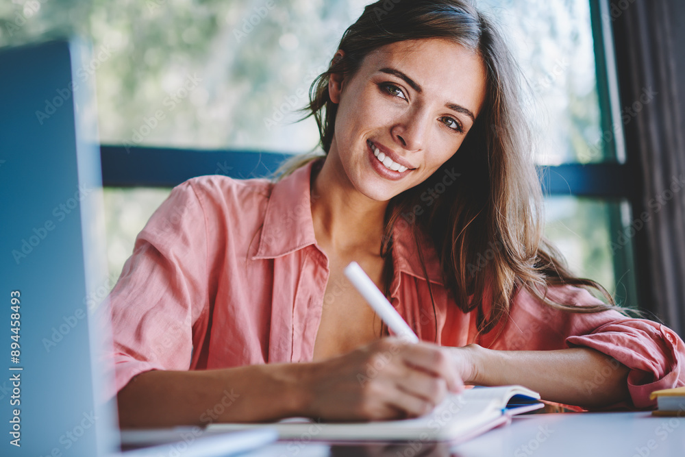 Poster portrait of successful young woman smiling at camera while writing down notes of working plan in not