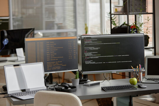 Background shot of empty programmer desk setup with laptop and multiple computer screens displaying source code programming at office workplace