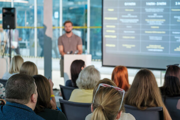 Business professional giving a presentation to an audience in a modern conference room setting with a large screen.