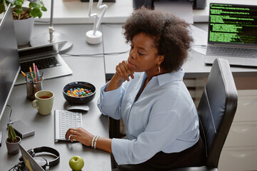 High-angle shot of thoughtful female African American senior IT specialist reading computer screen,...