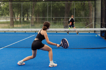 Two young players engaging in a competitive game of padel tennis on a blue outdoor court surrounded...