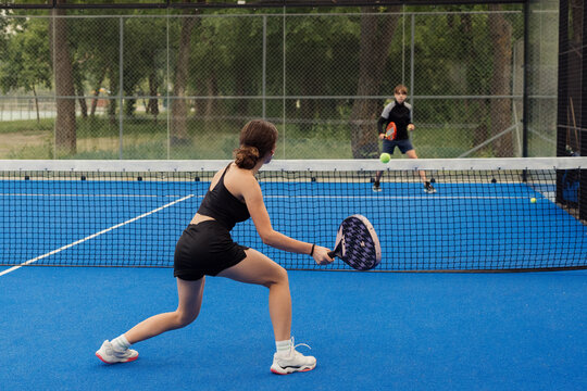 Two young players engaging in a competitive game of padel tennis on a blue outdoor court surrounded by a green park.