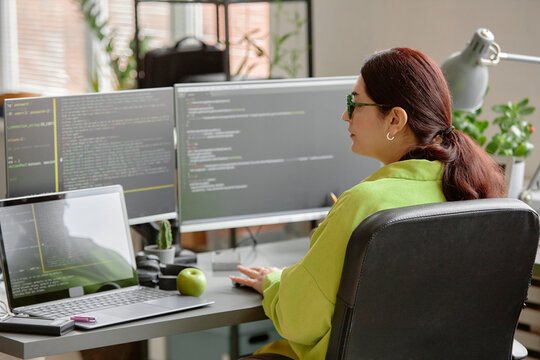 Rear view of young red-haired female programmer writing code at working desk with green apple in software development company office, copy space