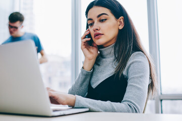 Serious businesswoman calling on smartphone while checking email on laptop computer using wireless internet.Pensive student talking on telephone during working at netbook sitting at desktop in office