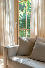 Fototapeta premium Close-up of a sofa in a living room with a window and curtains. The couch has a beige linen fabric. Light colors are used. Sunlight is coming through the trees on this sunny day.