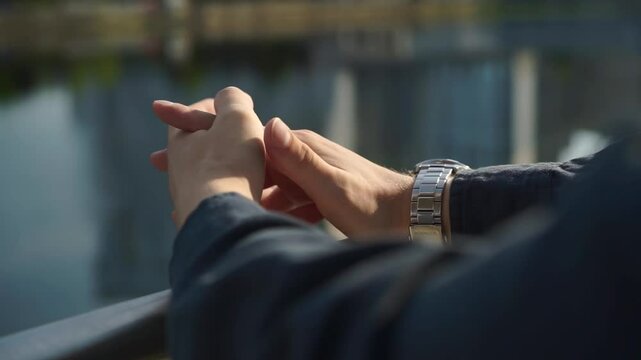 A man stands in a park by a lake, his hands show signs of mental illness, he fidgets nervously, itches and clenches his fists.