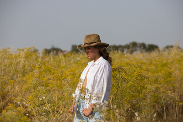 portrait of a beautiful young blonde woman standing in a field of oats. The woman is a farmer and wears a straw hat on her head. In the background the oat and fennel plantation. Organic farming.