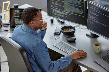 High-angle side view of thoughtful male African American software developer reading computer screen finding errors in code while sitting at desk with tea and candy bowl at hand in office, copy space