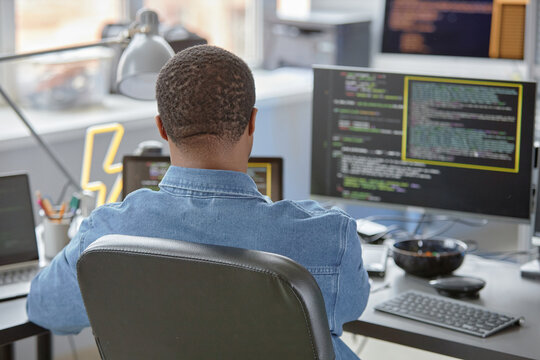 Rear view of male African American software engineer using computer at office desk for writing program code and finding errors