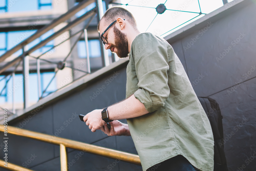 Poster casual dressed young man going up stairs and reading incoming notification on modern smartphone devi