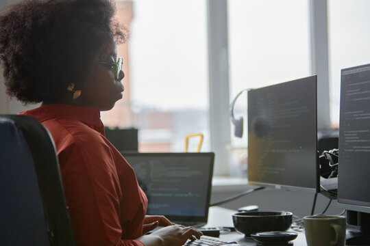 Side view of concentrated female African American IT engineer code programming on computer writing script while sitting at desk against window in office, copy space - Powered by Adobe