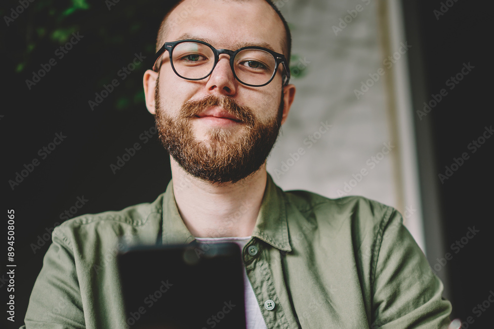 Poster below portrait of bearded hipster student in stylish eyeglasses for vision correction holding modern