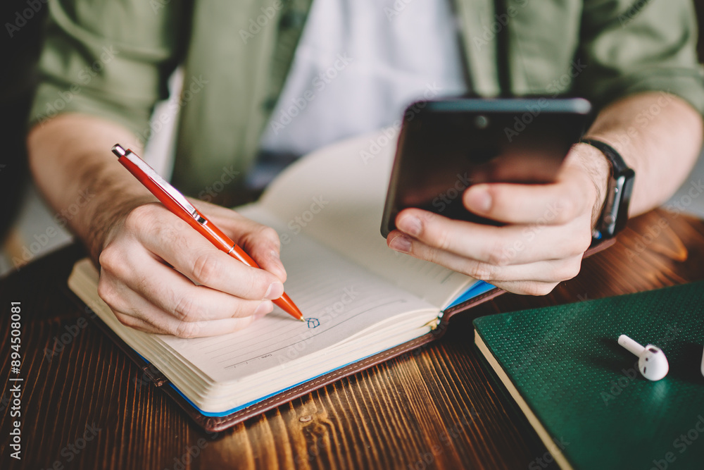 Poster cropped view of young man holding smartphone in hands and drawing sketch in notepad sitting at woode