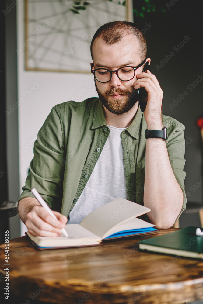 Poster pensive bearded young man calling on smartphone and writing down notes in notepad sitting at coffee 