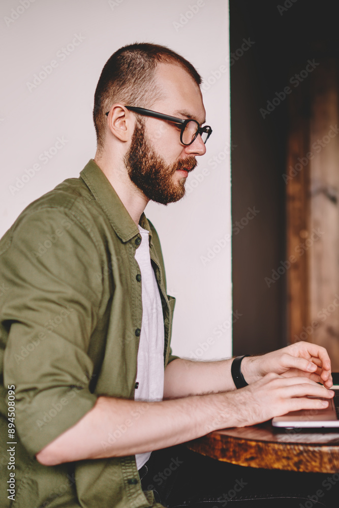 Poster side view of pensive young man freelancer typing information on keyboard of modern laptop device usi