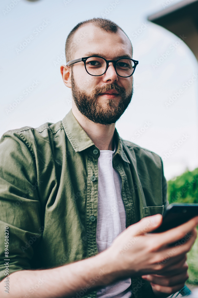 Poster portrait of bearded young man in eyeglasses holding telephone in hands and updating app using intern