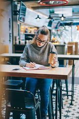 Concentrated female student sitting at cafeteria with cup of coffee in hand while writing organization plan, serious hipster girl casual dressed studying and notes main theses of day in textbook