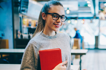 Half length portrait of cheerful female student standing near table at cafeteria and using mobile...