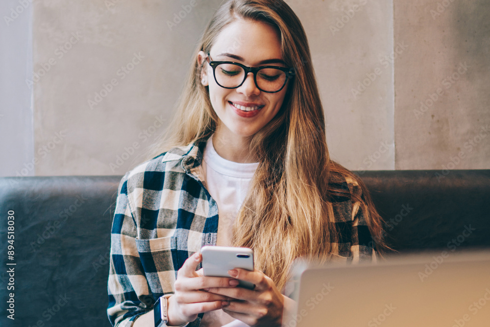 Poster smiling hipster girl messaging with friends via cellular phone after working on modern laptop comput