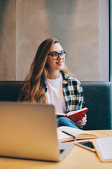 Positive smiling hipster girl wear in eyeglasses for vision correction spending time at coworking space reading book using computer and preparing for exam while looking away and waiting friend