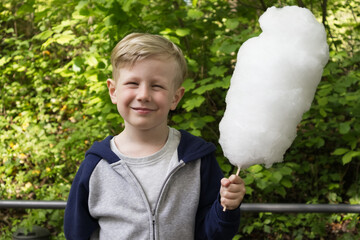 A boy holds a large white candy floss on a stick against a background of green foliage