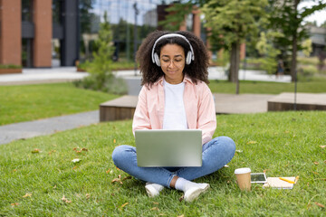 Young woman sitting on grass using laptop and wearing headphones. Outdoor study session in urban park. Fresh air and green surroundings creating a pleasant learning environment.