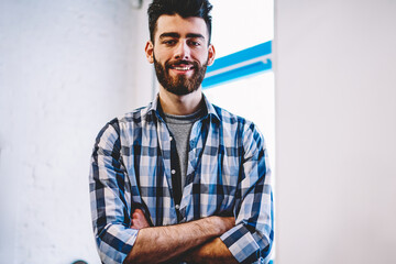 Half length portrait of successful male teenager looking at camera with cute smile on face at college campus, positive caucasian bearded hipster guy wear in trendy casual look enjoying time indoors