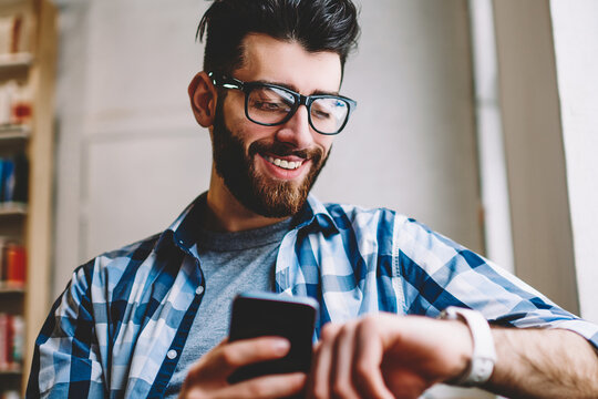 Smiling successful hipster guy in eyewear checking time on wearable smartwatch while waiting for meeting at university campus, happy man holding mobile phone in hand and reading message on wristwatch