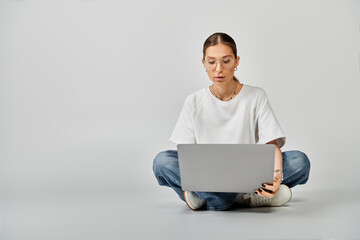 A young woman in a white t-shirt and glasses sitting on the floor, engrossed in her laptop on a grey background.