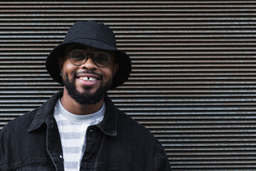 Waist up shot of happy black man smiles happily dressed in black hat and denim jacket being in good mood looks directly at camera expresses positive emotions