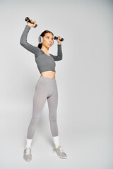 A sporty young woman in active wear is exercising with dumbbells on a grey background.