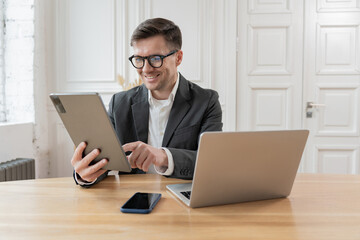 Smiling man in glasses and suit using a tablet, seated at a desk with a laptop and smartphone.