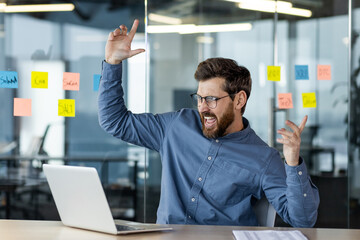 Excited man celebrating success in modern office near laptop with sticky notes in background